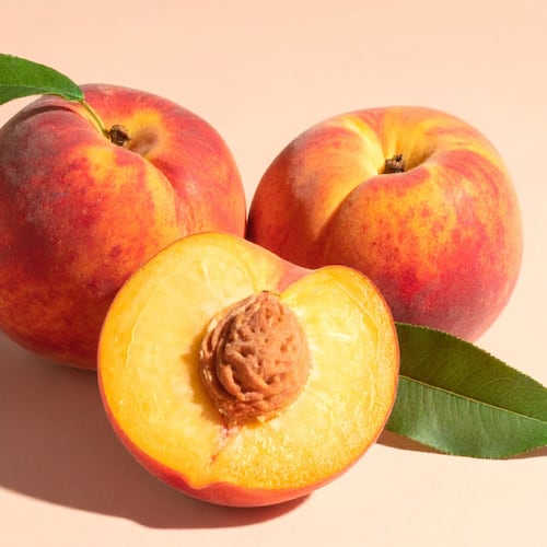Two whole peaches sit on a peach background with peach leaves still attached to them along with a sliced peach, showcasing the seed and bright yellow flesh.