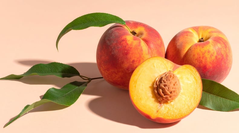 Two whole peaches sit on a peach background with peach leaves still attached to them along with a sliced peach, showcasing the seed and bright yellow flesh.