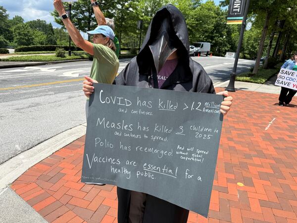 A costumed angel of death in a plague mask joined more than 50 protesters outside the Centers for Disease Control and Prevention in Atlanta on Tuesday, June 10, 2025, rallying against the newly released permanent health budget cuts in the Trump administration's budget.  The person wearing the costume did not want to give her name for fear of repercussions at her job, teaching in public health. But she said she is a Ph.D. and immunologist who previously was a researcher at the CDC. "I disagree with all of the choices people are making out of fear and ignorance," she said, which she said would lead to more death. Her sign spoke of the damage from COVID and measles and of the lifesaving value of vaccines. The plague mask was designed for plague doctors under the hope that the herbs stashed in the elongated beak nose could fight "bad air." (Ariel Hart/AJC)