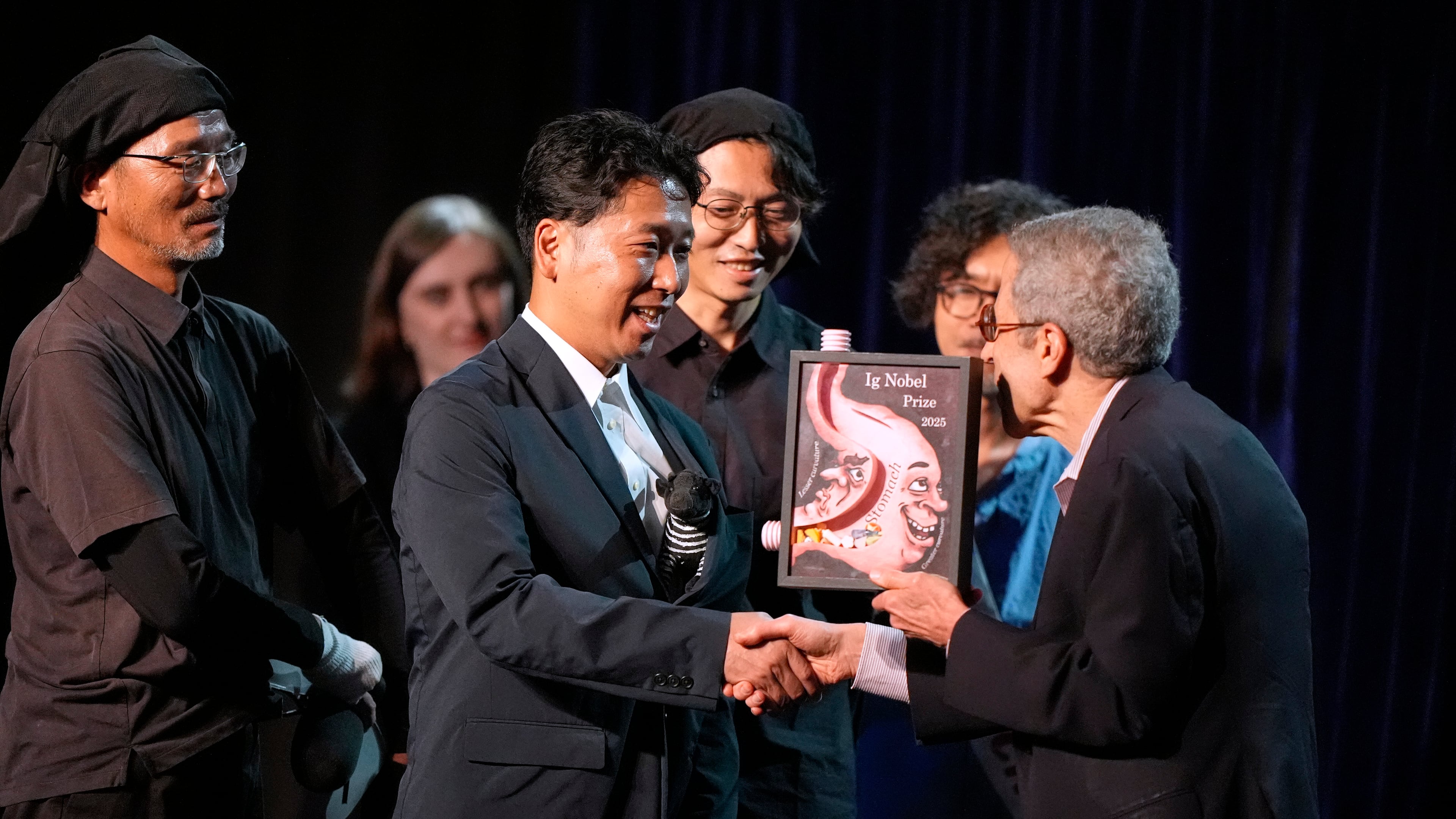 The Ig Nobel Prize for biology is accepted by Tomoki Kojima, Kazato Oishi, Yasushi Matsubara, Yuki Uchiyama, Yoshihiko Fukushima, Naoto Aoki, Say Sato, Tatsuaki Masuda, Junichi Ueda, Hiroyuki Hirooka, and Katsutoshi Kino, for their experiments to learn whether cows painted with zebra- like striping can avoid being bitten by flies, Thursday, Sept. 18, 2025, in Boston. (AP Photo/Robert F. Bukaty)