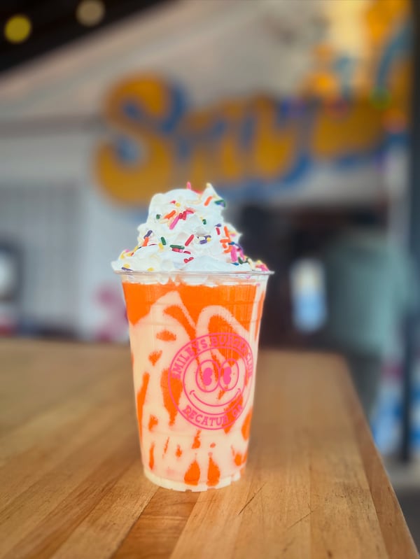 A frozen drink in a Smiley's Burger plastic cup is topped with whipped cream and colorful sprinkles. It sits on a wooden table, and a Smiley's Burger sign is out of focus in the background.