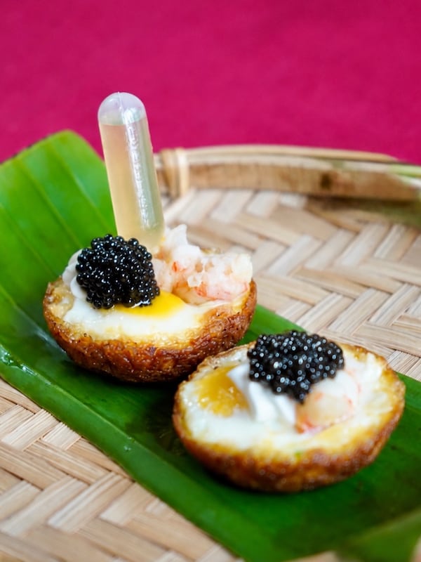 Two quail egg halves containing langostino  and topped with caviar are presented on a leaf, which is resting on a bamboo tray. There is a very small vial of fish sauce tucked into one of the egg halves.