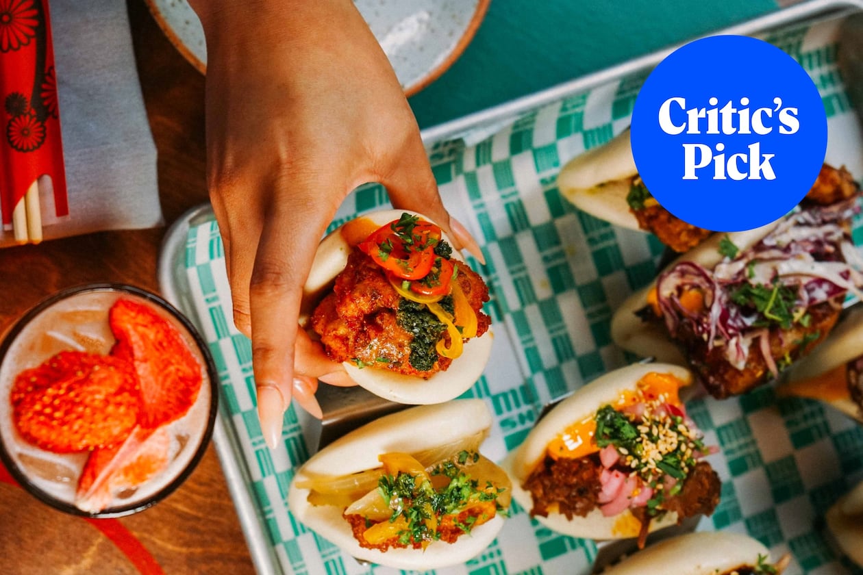 Overhead shot of several bao buns, including one that is being lifted by a hand. A strawberry highball cocktail sits to the left of the tray of buns