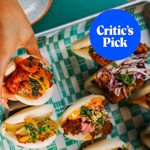 Overhead shot of several bao buns, including one that is being lifted by a hand. A strawberry highball cocktail sits to the left of the tray of buns