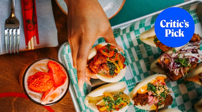 Overhead shot of several bao buns, including one that is being lifted by a hand. A strawberry highball cocktail sits to the left of the tray of buns