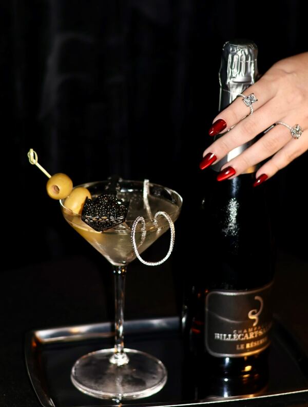 woman's jeweled hand holds a bottle of Champagne to the right of a martini garnished with olives, caviar and a diamond necklace.