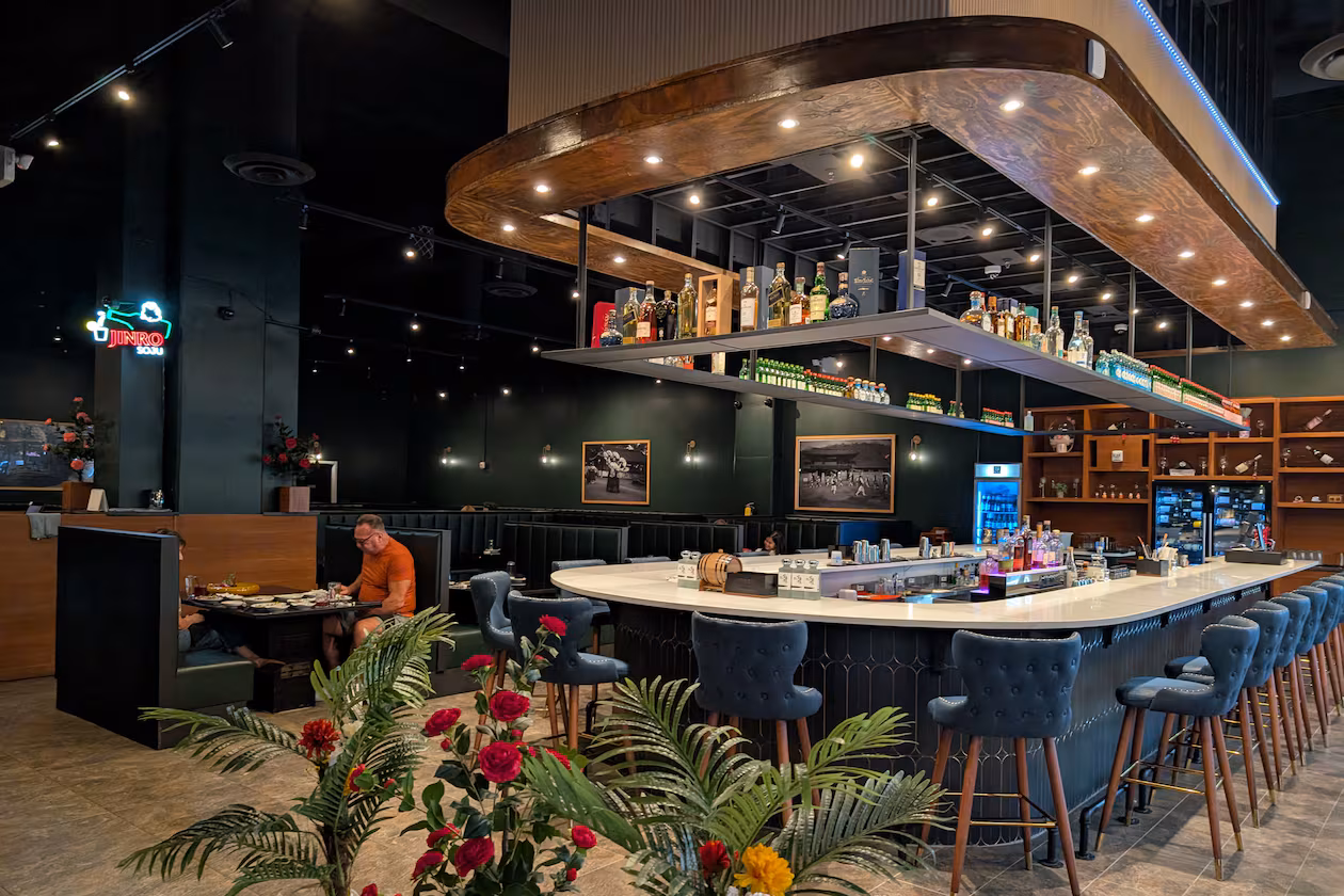 A long, modern, u-shaped restaurant bar sits in the middle of a restaruant dining room with lighting and shelves full of bottles overhead.