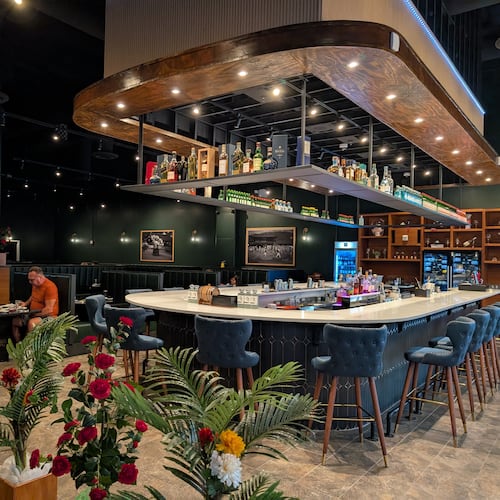 A long, modern, u-shaped restaurant bar sits in the middle of a restaruant dining room with lighting and shelves full of bottles overhead.