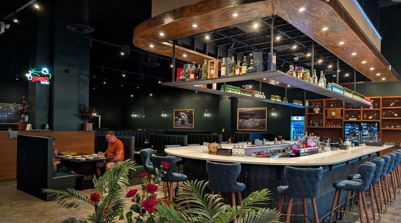 A long, modern, u-shaped restaurant bar sits in the middle of a restaruant dining room with lighting and shelves full of bottles overhead.