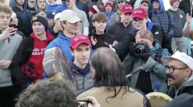 In this Friday, Jan. 18, 2019 image made from video provided by the Survival Media Agency, a teenager wearing a "Make America Great Again" hat, center left, stands in front of an elderly Native American singing and playing a drum in Washington. The Roman Catholic Diocese of Covington in Kentucky is looking into this and other videos that show youths, possibly from the diocese's all-male Covington Catholic High School, mocking Native Americans at a rally in Washington.