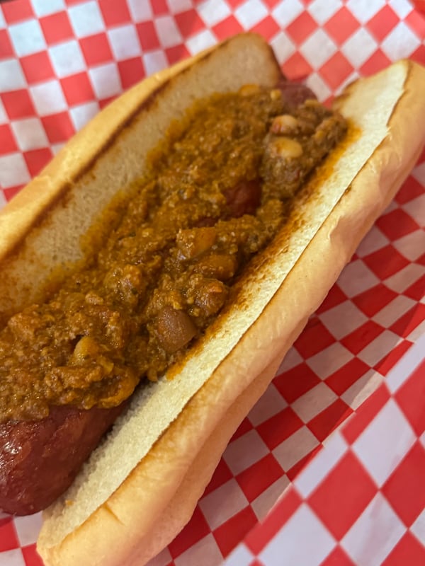 A chili dog in a buttered, grilled bun, placed on top of a red checkered paper wrapper.