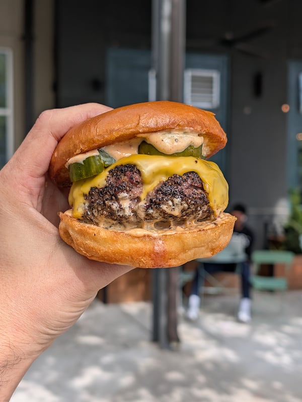 A hand holds an overly thick burger patty on a smooth bun. The burger is seasoned with peppercorns, and melted cheese drapes the top. It has pickle slices and a sauce on top of the patty as well.