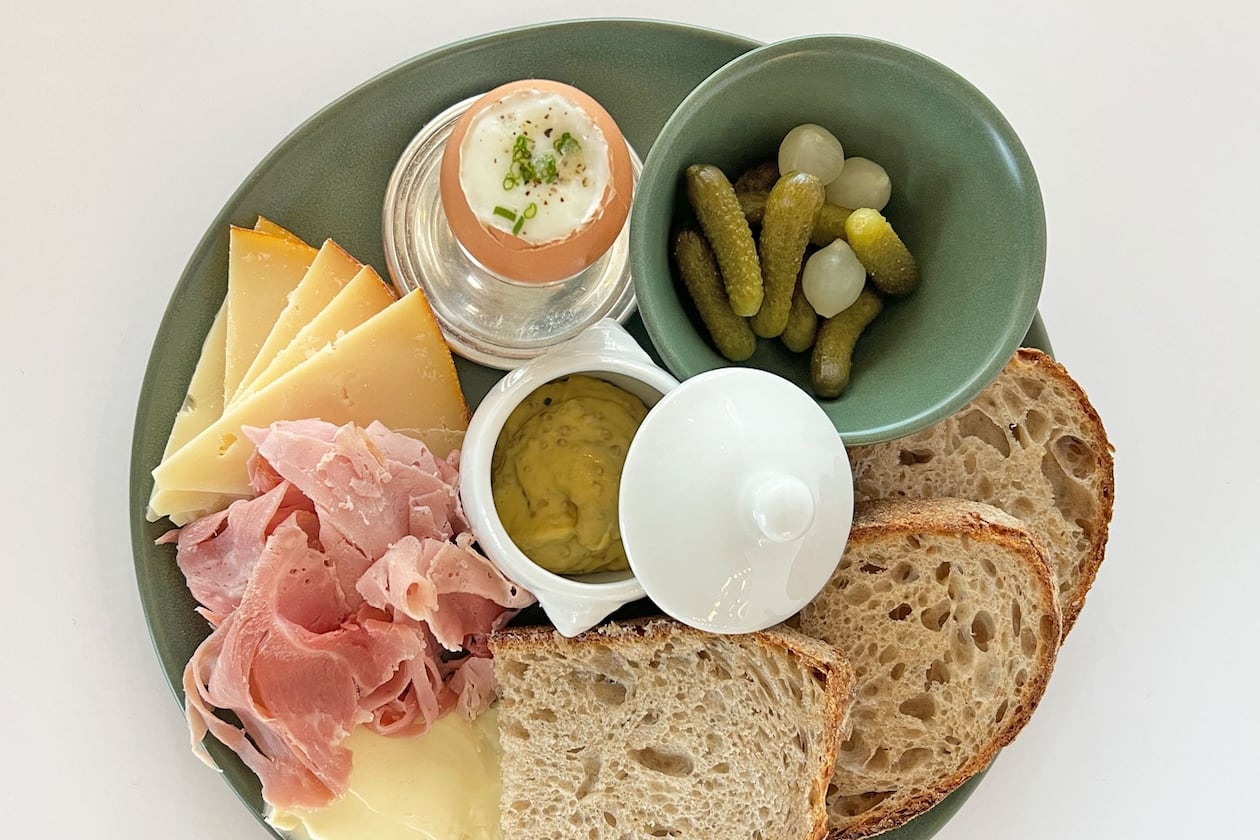 A plate at Summerland Cafe holds ham, various cheeses, a soft boiled egg, two slices of sourdough, a bowl of cornichon pickles and a pot of butter.