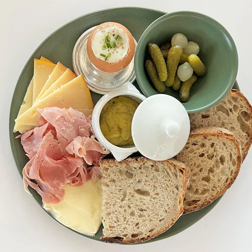 A plate at Summerland Cafe holds ham, various cheeses, a soft boiled egg, two slices of sourdough, a bowl of cornichon pickles and a pot of butter.