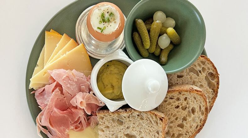 A plate at Summerland Cafe holds ham, various cheeses, a soft boiled egg, two slices of sourdough, a bowl of cornichon pickles and a pot of butter.