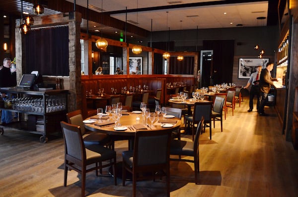 The dimly lit interior of Oak Steakhouse. The dining room has round tables for groups and banquet seating as well. Restaurant staff is well-dressed, with one person wearing a tie and holding a tray, and a man wearing a sports coat standing at the bar.