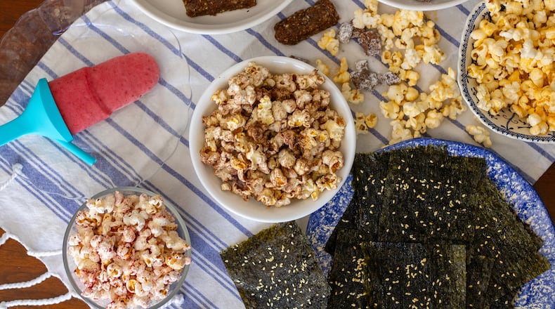 Making snacks with kids is a fun activity for all ages. From left, clockwise: Raspberry-Mango Smoothie Pop, Cherry and Walnut Snack Bars, Sweet and Savory Puppy Chow, cheese-flavored popcorn, Sesame Seaweed Snacks, freeze-dried fruit-flavored popcorn and hot cocoa mix-flavored popcorn (center). (Kate Williams for The Atlanta Journal-Constitution)