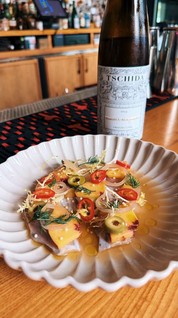 A plated trout crudo garnished with sliced peaches, peppers, olives, and greens, served alongside a bottle of Christian Tschida Gruner Veltliner wine on a bar counter.
