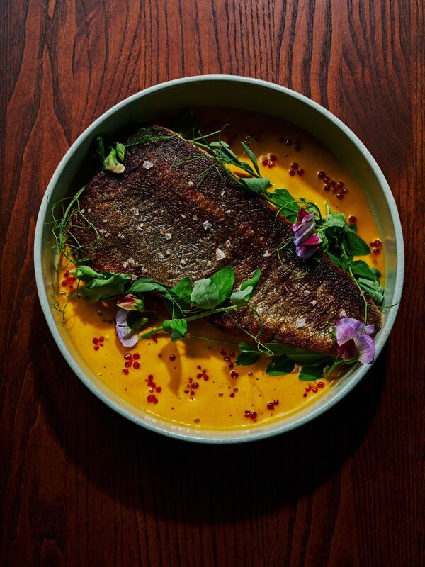 A top-down view of a dish of trout with the skin side up over an orange sauce and garnished with green herbs and purple flowers