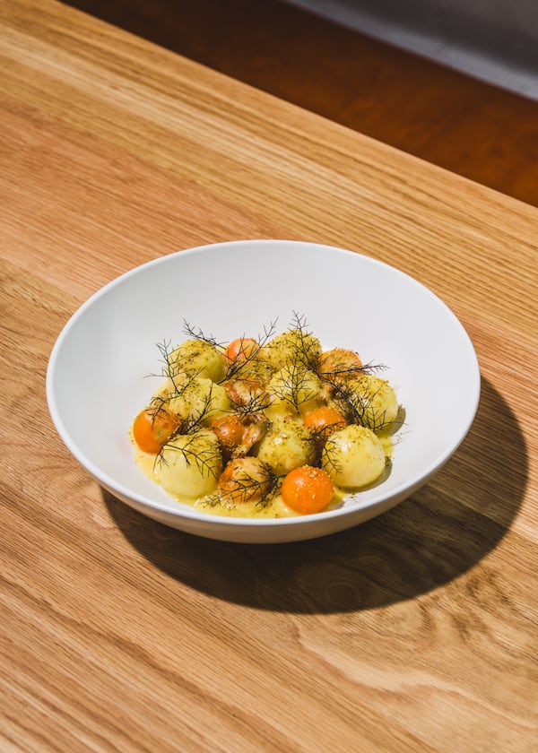 A bright white bowl of corn dumplings interspersed with sungold tomatoes, all roughly the size of cherries, on top of a corn sauce with springs of herbs sticking up from the top.