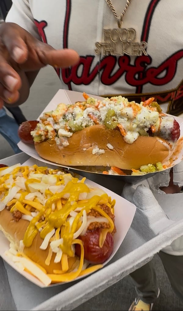 A man in a Braves jersey extends two hot dogs loaded with toppings toward the camera.