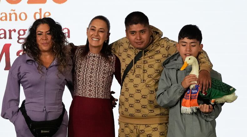 Merlin the duck, dressed in a Mexican national soccer team jersey, attends the daily press conference by Mexican President Claudia Sheinbaum, second from left, along with his caretakers Carla Gomez and her sons Carlos, and Christian who holds Merlin, at the National Palace in Mexico City, Monday, June 22, 2026. (AP Photo/Marco Ugarte)