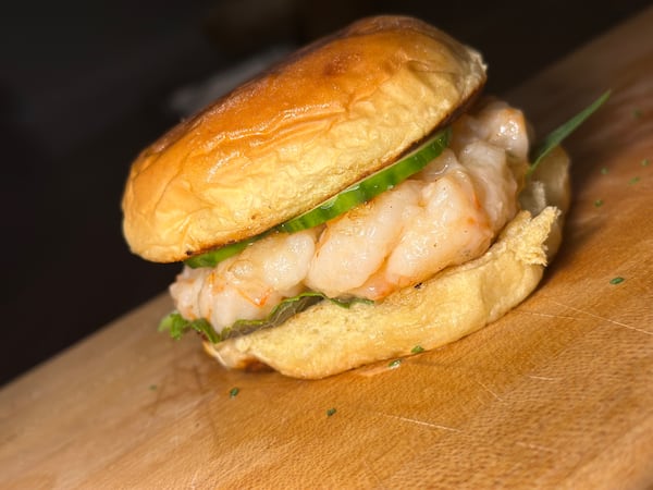 A brioche bun filled with shrimp is placed on a wood-topped table. The burger is garnished with lettuce. The photo is taken at an angle, so the shrimp burger is tilted.
