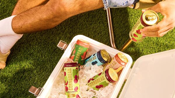 A top-down shot shows a cooler filled with ice and cans of Cornbread beverages. Visible are a man's legs and his hand holding one of the cans; he appears to be sitting in a lawn chair next to the cooler.