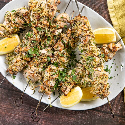 Six charred chicken kebabs on metal skewers are arranged on a serving platter placed on a wood tabletop. The kebabs are generously sprinkled with chopped Italian parsley leaves, with lemon wedges on the side. A lemon-yellow towel is arranged next to the serving platter.