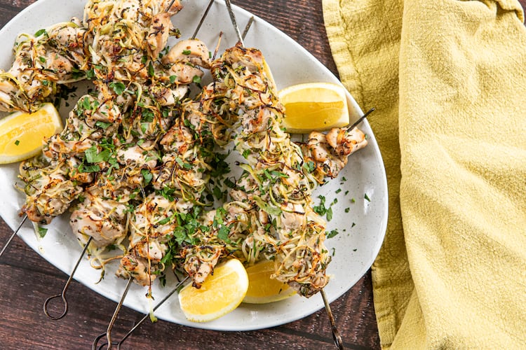 Six charred chicken kebabs on metal skewers are arranged on a serving platter placed on a wood tabletop. The kebabs are generously sprinkled with chopped Italian parsley leaves, with lemon wedges on the side. A lemon-yellow towel is arranged next to the serving platter.