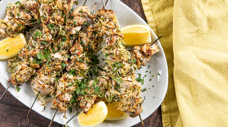 Six charred chicken kebabs on metal skewers are arranged on a serving platter placed on a wood tabletop. The kebabs are generously sprinkled with chopped Italian parsley leaves, with lemon wedges on the side. A lemon-yellow towel is arranged next to the serving platter.