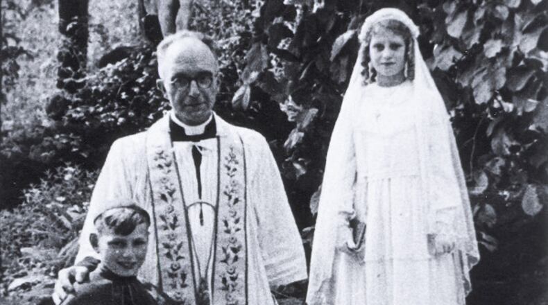 Holocaust survivor Beatrice Muchman, while in hiding, wearing her first communion dress. She is standing next to Father Vaes and her cousin, Henri, a choir boy.
