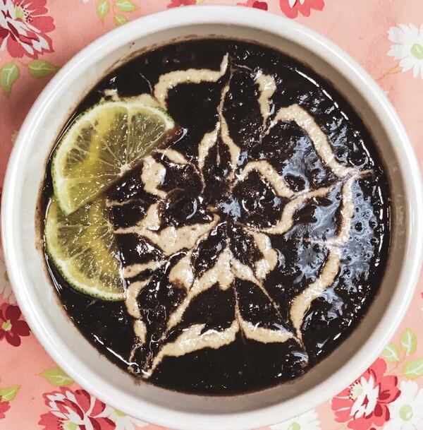 A bowl of black bean soup is garnished with a drizzle of cashew cream that resembles a flower. It is garnished with two slivers of lemon peel.