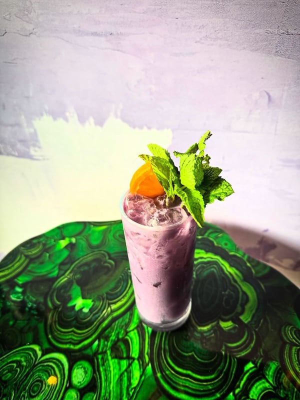 A vibrant, colorful photo showing a pastel milky cocktail in a highball glass, garnished with mint leaves and an orange wedge.