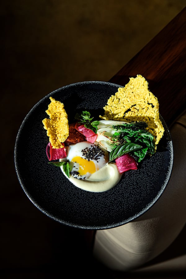 A highly styled photo shows a poached egg on a dark plate surrounded by colorful garnishes, including grilled bok choy.