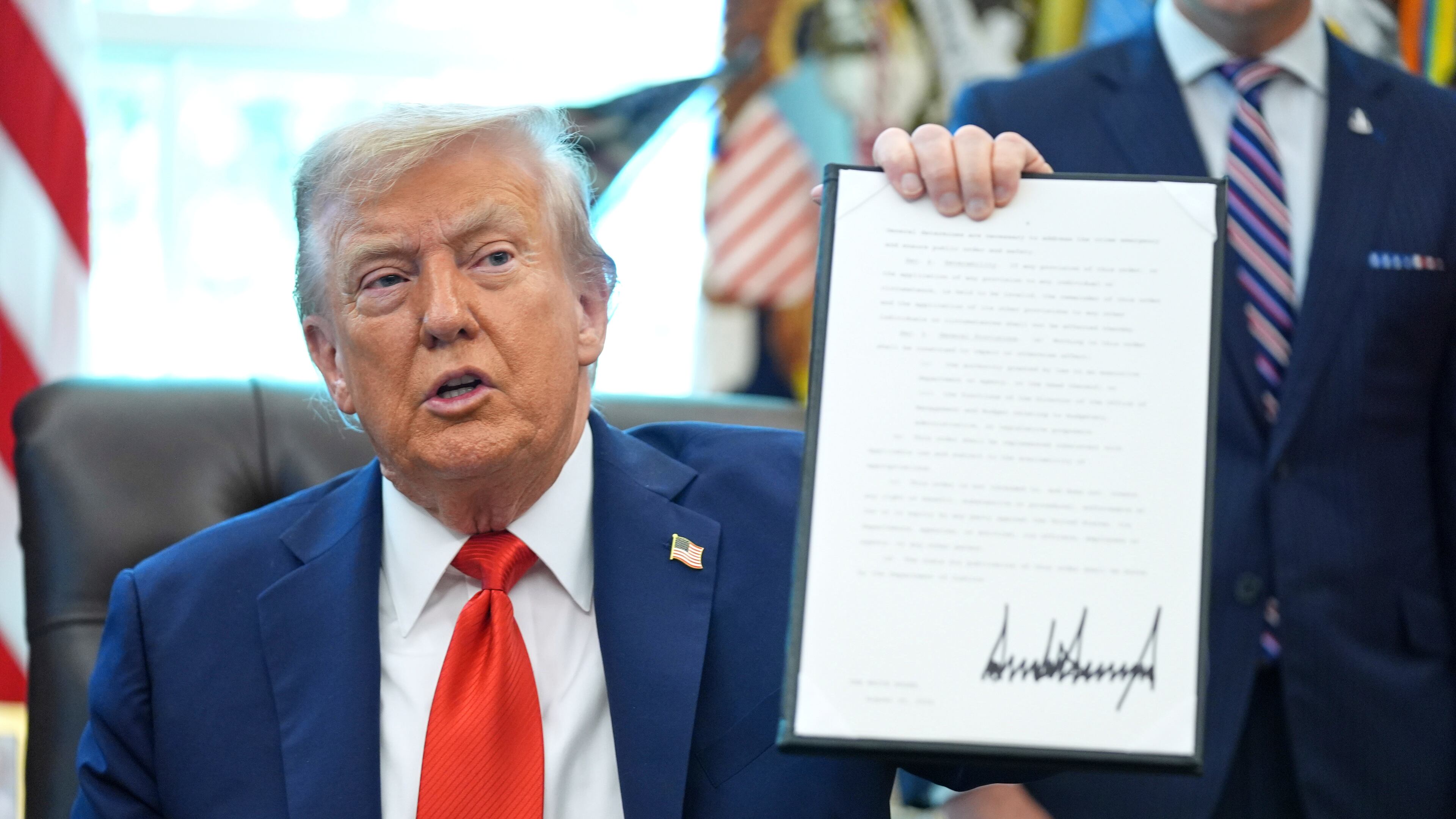 President Donald Trump holds up a signed executive order requiring the Justice Department to investigate instances of flag burning, in the Oval Office of the White House, Monday, Aug. 25, 2025, in Washington. (AP Photo/Evan Vucci)