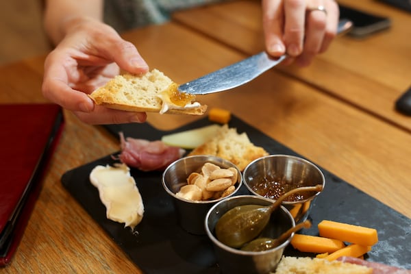 A cheese board is angled on a wood-topped table. The board contains a few pieces of yellow and white cheese, and containers of nuts, jam and pepperocini. Over the board, the hands of a person using a kife to smear jam onto a piece of bread are shown.