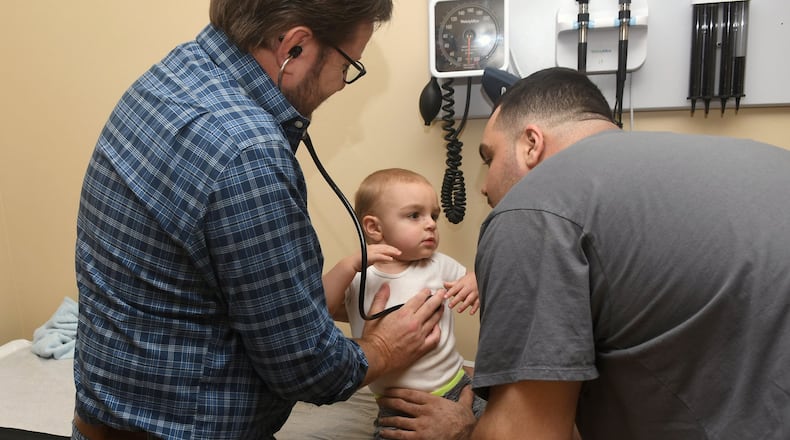 Dr. Greg McHan, in the checked shirt and jeans, examines Lenny Falletta III, 18 months, of Blairsville with a little help from Lenny Falletta Jr., his dad. They are at the Georgia Mountains Health clinic in Blue Ridge. Georgia Mountains Health needs to add another pediatrician, which is not easy in a rural area. But as Congress let the national clinic program’s funding expire, Georgia Mountains has put the brakes on attending recruiting fairs and hiring a recruitment firm. CEO Steve Miracle said he “can’t in good conscience” say federal funding will be there to sign a multiyear contract. (Photo by Rebecca Breyer / special to the AJC)