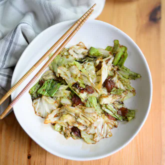 A bowl of stir-fried cabbage with bacon and sauce. Chopsticks rest on the bowl, and a dish towel rests against the bowl.