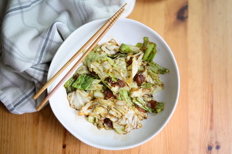 A bowl of stir-fried cabbage with bacon and sauce. Chopsticks rest on the bowl, and a dish towel rests against the bowl.