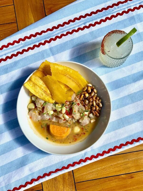 A bowl of ceviche from Tio Lucho's sits on a blue and white striped tablecloth with a cocktail beside it.