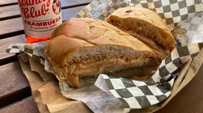 NYC Bodega offers a variety of handhelds from empanadas to Cuban sandwiches to chimi burgers (pictured). The latter is a Dominican street food favorite featuring a beef patty topped with shredded cabbage, sliced tomato and a slather of mayo-ketchup sauce served on bread known as pan de agua. (Ligaya Figueras / ligaya.figueras@ajc.com)