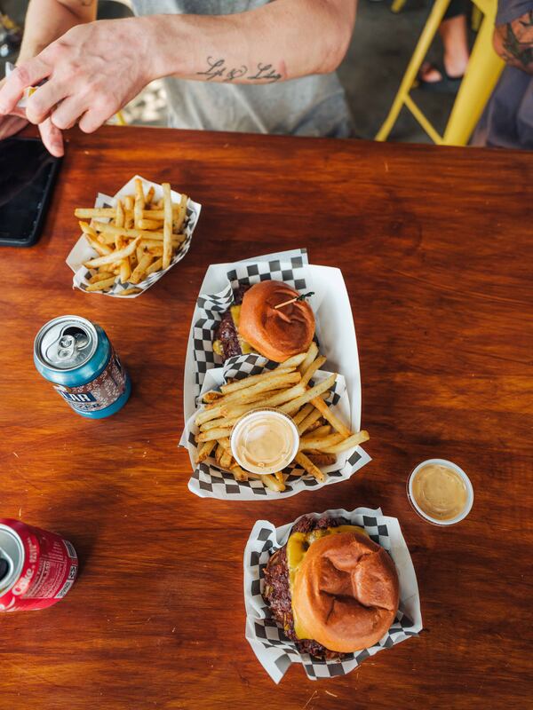 Overhead shot of a restaurant table with a couple of burgers and a couple of sides of fries, dipping sauces and sodas.