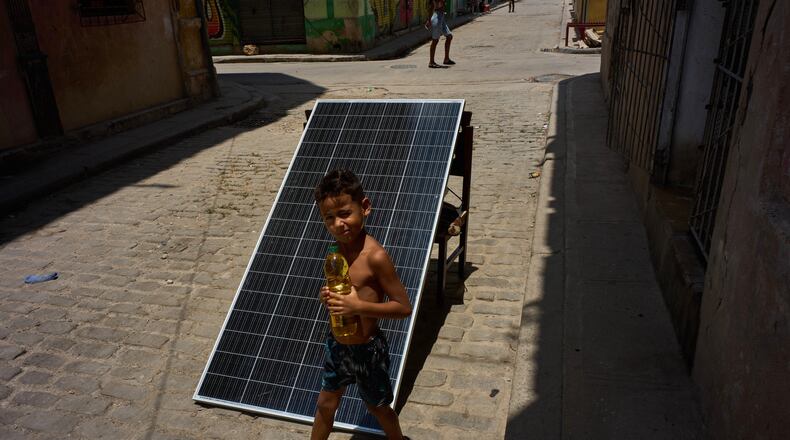 A child walks with a bottle of oil past a solar panel set up on the street to charge batteries during a blackout in Havana, Cuba, Monday, July 6, 2026. (AP Photo/Ramon Espinosa)