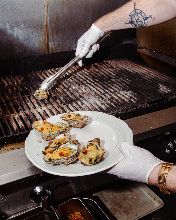 A person wearing white gloves uses metal tongs to transfer grilled oysters topped with melted cheese and diced bacon from a large grill grate onto a white plate.