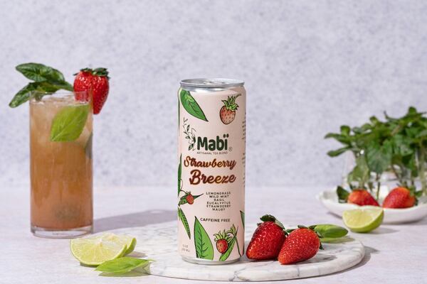 A can of Mabi Strawberry Breeze cold brew tea is placed on a marble stone, accompanied by a glass jar filled with the tea and garnished with a strawberry, lime wedges and a green herb leaf. More of the garnishes are also plated in the photo.