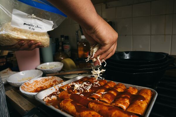 One hand holds a zip-close bag of shredded cheese while the other hand sprinkles the cheese over a sheet pan filled with enchiladas covered in red sauce. The sheet pan appears to be on top of a residential stove. There are dishes and cast-iron skillets around the pan, indicating a busy home kitchen.