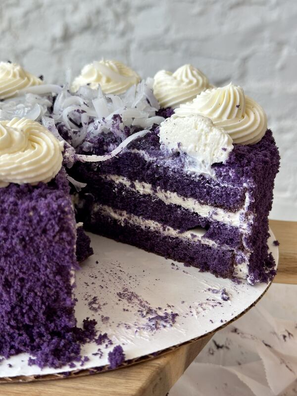 A close-up, sliced view of a layered purple ube cake topped with white whipped frosting and shredded coconut.