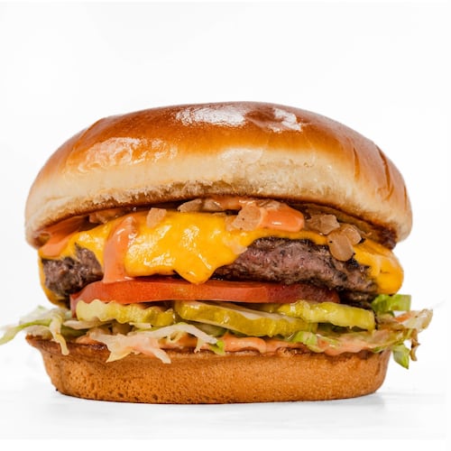 A classic-looking cheeseburger on a smooth, shiny bun, garnished with pickles, tomato slices, lettuce and a sauce.