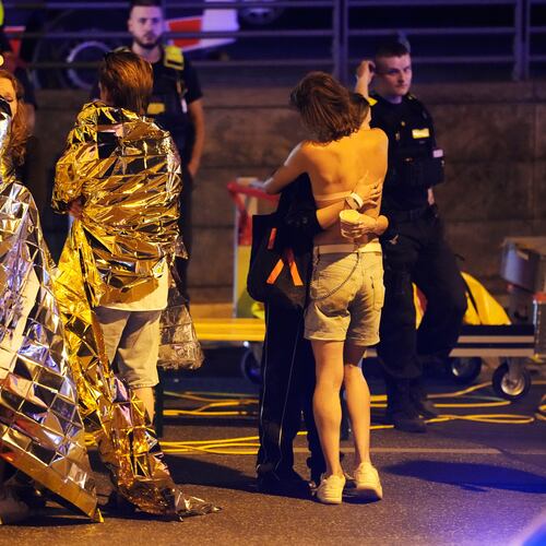 Two people embrace near others wearing thermal blankets as they stand near emergency personnel in Berlin, early Sunday, July 26, 2026, following the cancellation of Christopher Street Day festivities. (AP Photo/Markus Schreiber)